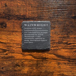 May include: A square, dark gray slate coaster with the word "WAINWRIGHT" and its pronunciation etched in white. The coaster also includes a definition and description. The coaster sits on a wooden surface.