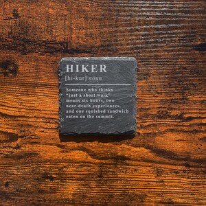 May include: A square, dark gray slate coaster with the definition of "HIKER" in white text. The definition includes the pronunciation, and a humorous description of what a hiker experiences. The coaster is on a wooden surface.
