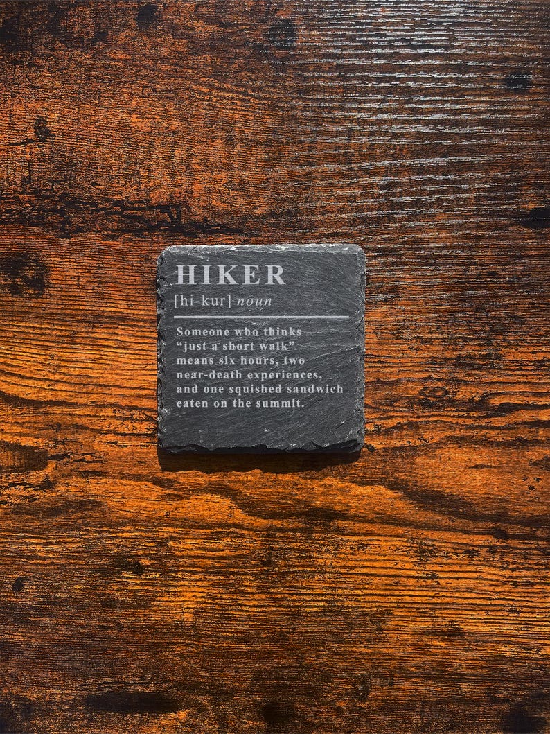 May include: A square, dark gray slate coaster with the definition of "HIKER" in white text. The definition includes the pronunciation, and a humorous description of what a hiker experiences. The coaster is on a wooden surface.