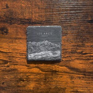 May include: A black slate coaster with a white line drawing of a mountain range and the text "LES ARCS FRANCE".