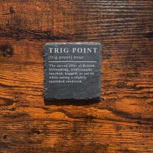 May include: A square, dark gray slate coaster with the text "TRIG POINT" and a definition. The coaster sits on a wooden surface with a warm, brown tone. The text describes a British hillwalking tradition.