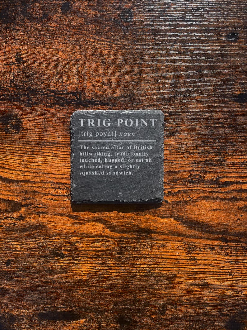 May include: A square, dark gray slate coaster with the text "TRIG POINT" and a definition. The coaster sits on a wooden surface with a warm, brown tone. The text describes a British hillwalking tradition.