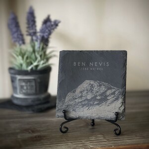 May include: A black slate plaque with a white engraved image of Ben Nevis, the highest mountain in the British Isles, and the text "Ben Nevis 1345 Metres". The plaque is on a black metal stand.