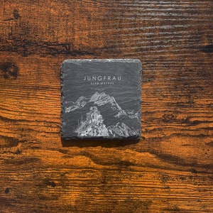 May include: A black slate coaster with a white outline of the Jungfrau mountain in Switzerland. The text "Jungfrau 4158 metres" is printed on the coaster.