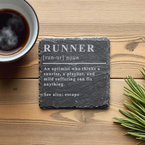 Puede incluir: Un posavasos de pizarra gris oscuro con la definición de "RUNNER" grabada en blanco. La definición incluye la pronunciación, una descripción y "Ver también: escape". Una taza de café está cerca sobre una superficie de madera.