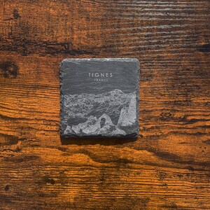 May include: A black slate coaster with a white etched image of a mountain range and the text "Tignes France".