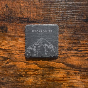 May include: A black slate coaster featuring a white outline of Dhaulagiri mountain and the text "DHAULAGIRI 8167 METRES" on a brown wooden surface.
