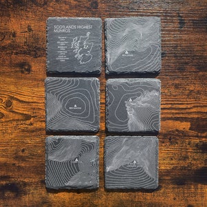 May include: Set of six black slate coasters featuring topographic maps of Scotland's highest mountains. Each coaster has a white outline of a mountain range and the name of the mountain, including Ben Nevis, Ben Macdui, Sgor an Lochain, Cairn Toul, and Braeriach.