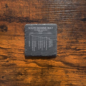 Peut inclure: Un dessous de verre en ardoise noire avec un graphique blanc de la carte du sentier South Downs Way. La carte montre les 100 miles du sentier de Winchester à Eastbourne, en Angleterre. Le nom du sentier, la distance et les points de départ et d'arrivée sont imprimés sur le dessous de verre.