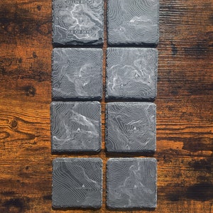 Puede incluir: Conjunto de seis posavasos de pizarra negra con mapas topográficos de la isla de Skye, Escocia. Cada posavasos presenta una cordillera diferente con el nombre de la montaña impreso en blanco.