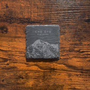May include: A black slate coaster with a white outline of a mountain range and the text "CHO OYU 8188 METRES".