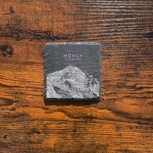 May include: A black slate coaster with a white engraved image of the Mönch mountain in Switzerland. The text "Mönch 4110 metres" is also engraved on the coaster.