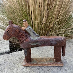 May include: A rustic ceramic sculpture of a horse with a rider, featuring a textured, dark reddish-brown finish. The horse has a blocky, stylised form with a rectangular base, while the rider is depicted in a lighter, muted grey-green tone.