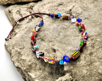 May include: A colorful beaded bracelet with a brown cord. The beads are various shapes and sizes, including round, rectangular, and oval, and feature a variety of colors and patterns. The bracelet is displayed on a gray stone surface.