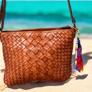 May include: A brown leather crossbody bag with a woven design. The bag has a long, thin strap and a small, colorful charm attached to the side. The background shows a sandy beach and the ocean.