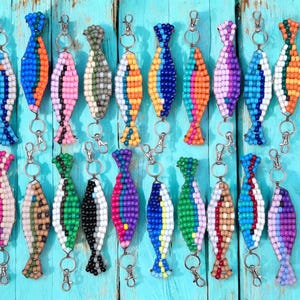 Puede incluir: Una colección de llaveros de peces con cuentas de varios colores, incluyendo azul, naranja, rosa, verde y morado. Cada llavero tiene un cierre plateado. Los llaveros están dispuestos sobre una superficie de madera turquesa.