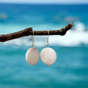 May include: Two circular, off-white earrings with a pearlescent finish, suspended from silver hooks. They hang from a brown branch, set against a blurred blue sea background. Each earring features a small silver bead at the base.
