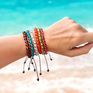 May include: A collection of colorful beaded bracelets in shades of brown, teal, white, orange, green, and pink. Each bracelet is strung on a dark cord with a sliding knot closure. The bracelets are displayed on a person's wrist against a backdrop of a beach.