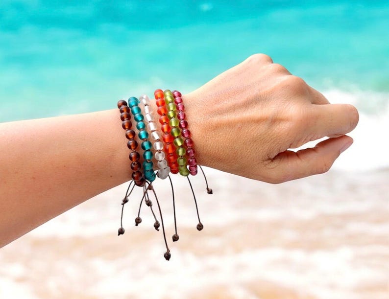 May include: A collection of colorful beaded bracelets in shades of brown, teal, white, orange, green, and pink. Each bracelet is strung on a dark cord with a sliding knot closure. The bracelets are displayed on a person's wrist against a backdrop of a beach.