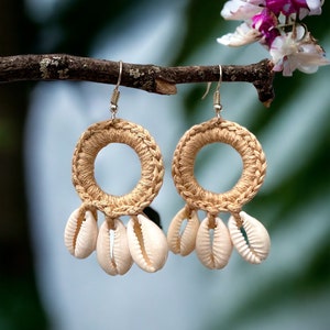 Könnte beinhalten: Handgefertigte Ohrringe mit einem geflochtenen, beigefarbenen Reifen-Design und herabhängenden Muscheln. Die Ohrringe hängen an silberfarbenen Haken und erzeugen einen natürlichen, strandähnlichen Look. Der Hintergrund ist verschwommen, mit grünen und rosa Farbtupfern.