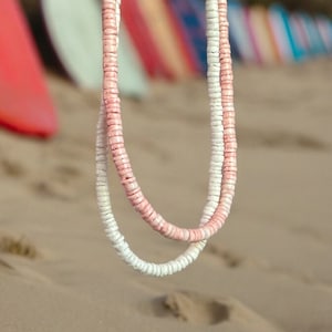 May include: Two layered necklaces, one white and one white with pink accents, made of small, round beads. The necklaces have a beachy aesthetic. Surfboards are visible in the background, enhancing the coastal theme. The necklaces are perfect for summer.