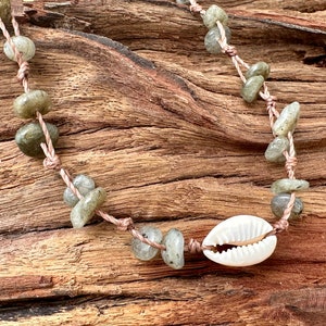 May include: A handcrafted necklace with pale green gemstone beads and a white seashell pendant. The beads are threaded on a light brown cord, giving a natural, earthy look. The necklace is shown on a wooden surface.