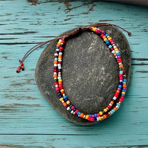 May include: A multi-coloured beaded anklet with a brown cord, displayed on a grey stone against a turquoise background. The anklet has rows of small, vibrant beads in red, orange, yellow, blue, and green, with silver accents.