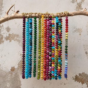 May include: A collection of eleven beaded necklaces hanging from a wooden branch. Ten necklaces feature uniform, solid-colored beads in shades of deep purple, blue, green, yellow, red, and pink. One necklace displays a multi-colored, rainbow-like pattern.