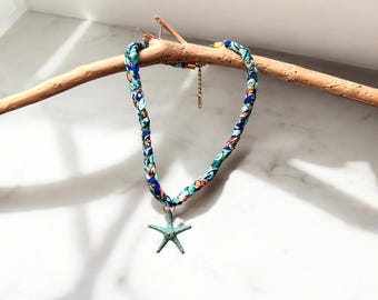 May include: A braided fabric necklace with a blue, green, and orange floral pattern. The necklace features a turquoise starfish pendant. The necklace is displayed on a wooden branch against a white background.