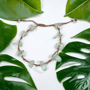 Könnte beinhalten: Ein handgefertigtes Armband mit hellgrünen Seeglasperlen, auf einer braunen Kordel aufgefädelt. Das verstellbare Armband enthält kleine silberfarbene Perlen. Das Design ist schlicht und elegant, geeignet für den Alltag.