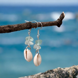 May include: Dangle earrings with natural seashells and sea glass beads. The earrings hang from silver-coloured hooks. The background shows a blurred view of the ocean and sky, creating a coastal theme.