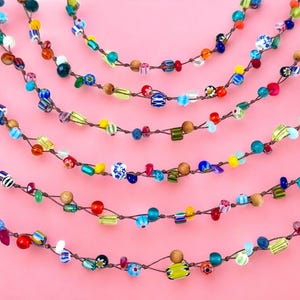 May include: Several strands of colorful beaded necklaces are displayed against a pink background. The beads are various shapes and sizes, including round, square, and cylindrical, in shades of blue, green, yellow, red, and white. The necklaces are strung on a brown cord.
