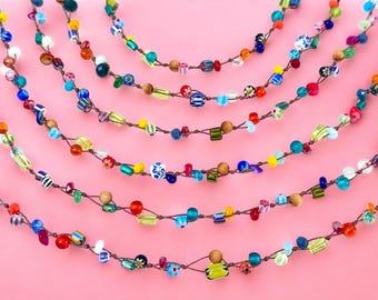 May include: Several strands of colorful beaded necklaces are displayed against a pink background. The beads are various shapes and sizes, including round, square, and cylindrical, in shades of blue, green, yellow, red, and white. The necklaces are strung on a brown cord.