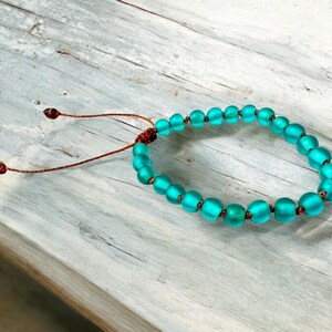 May include: A turquoise beaded bracelet with a sliding knot closure. The bracelet features round, frosted glass beads in varying sizes, strung on a brown cord. The beads are a vibrant teal color, and the bracelet is displayed on a weathered wooden surface.