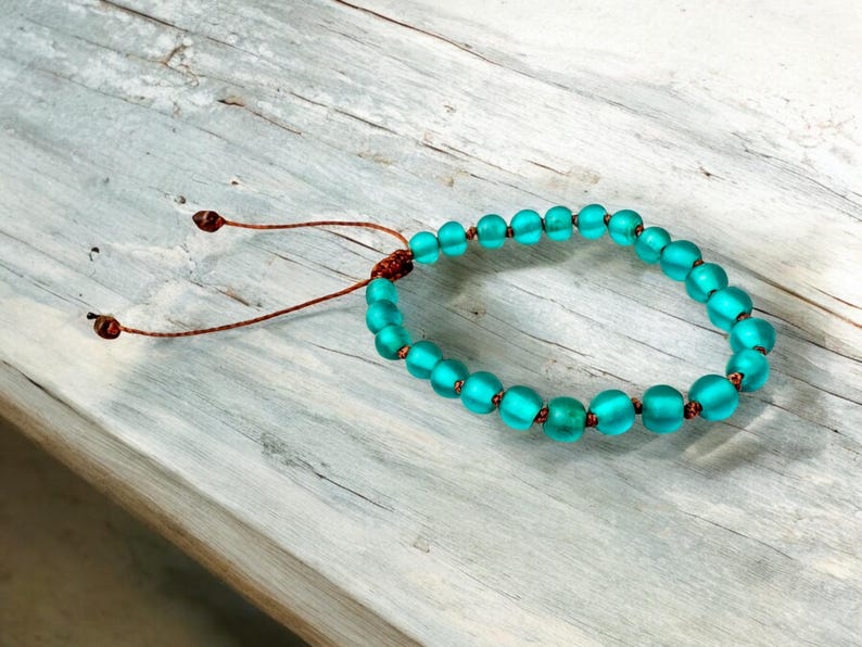 May include: A turquoise beaded bracelet with a sliding knot closure. The bracelet features round, frosted glass beads in varying sizes, strung on a brown cord. The beads are a vibrant teal color, and the bracelet is displayed on a weathered wooden surface.