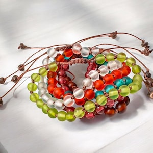 May include: A collection of colorful beaded bracelets. The bracelets feature translucent glass beads in shades of red, orange, green, white, and teal. Each bracelet is strung on a brown cord with decorative knots at the ends.