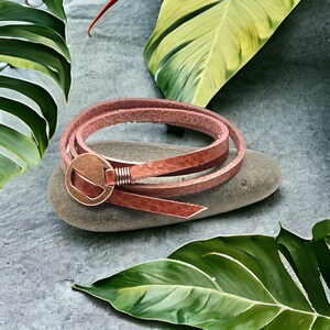 May include: A wrap-around bracelet in brown leather, featuring a copper-coloured clasp. The bracelet is coiled around a smooth, grey stone. Green leaves are in the background.