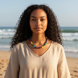 May include: A woman wearing a beige V-neck top and a colourful beaded necklace stands on a sandy beach with the sea in the background. The necklace features a variety of coloured beads, including red, yellow, and blue.