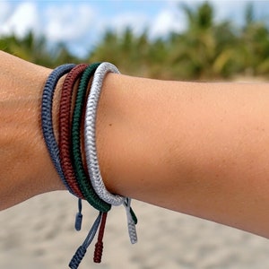 May include: A collection of braided rope bracelets in various colors, including blue, burgundy, green, and silver. Each bracelet has adjustable ties. The bracelets are shown on a person's wrist, with a beach and palm trees in the background.