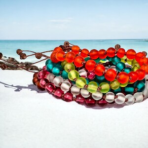 May include: A stack of colorful beaded bracelets in shades of red, orange, green, teal, and clear. The bracelets are on a white surface with a beach and ocean background.