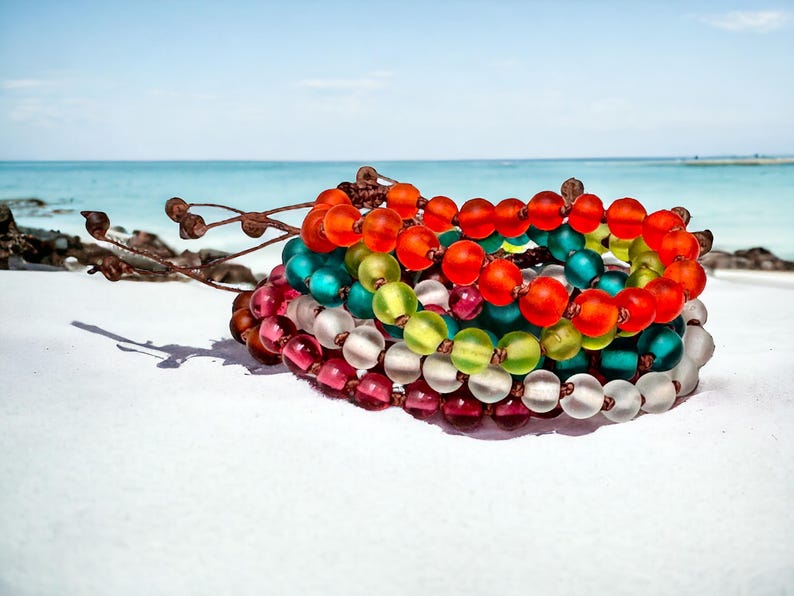 May include: A stack of colorful beaded bracelets in shades of red, orange, green, teal, and clear. The bracelets are on a white surface with a beach and ocean background.