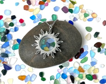 May include: A silver sun-shaped pendant necklace with a clear center filled with colorful sea glass, resting on a dark gray stone. The necklace is on a silver chain. Colorful sea glass pieces are scattered around the stone.