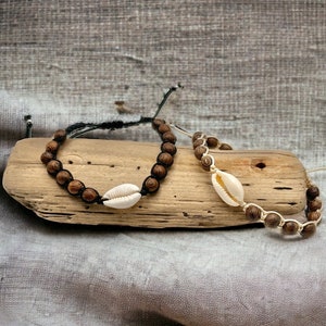 May include: Two handmade bracelets. One features dark brown wooden beads and a white seashell on a black cord. The other bracelet has light brown wooden beads and a white seashell on a cream-colored cord. Both are displayed on a piece of driftwood.