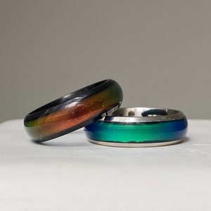 Peut inclure: Deux bagues d'humeur avec des anneaux à changement de couleur. Une bague présente une finition en métal noir avec un dégradé de teintes orange, vert et jaune. La seconde bague a une finition en métal argenté avec une surface dégradée vert et bleu.