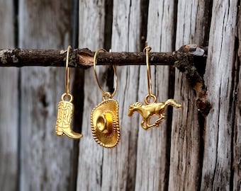 May include: Gold-colored earrings featuring a cowboy boot, a cowboy hat, and a running horse charm. The earrings are hanging from a small branch against a weathered wood background. The charms are detailed and shiny.