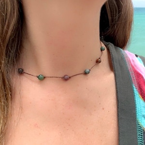 May include: A choker necklace with small, multi-coloured beads in green, brown, and purple. The beads are strung on a thin, brown cord. The necklace sits close to the neck. The background shows a glimpse of the sea.