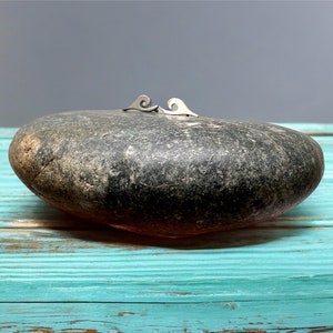 May include: A pair of silver wave-shaped earrings. The earrings are displayed on a large, dark gray stone, set against a turquoise wooden surface and a gray background. The earrings are small and delicate, with a minimalist design.