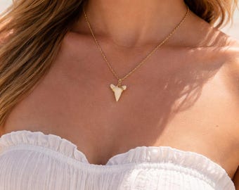 May include: A gold-colored necklace with a shark tooth pendant. The pendant is white and triangular, suspended from a delicate chain. The model is wearing a white strapless top.