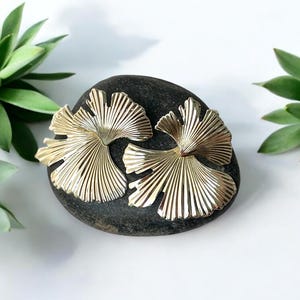 Puede incluir: Dos pendientes dorados en forma de abanico descansan sobre una piedra gris oscura. Los pendientes tienen un diseño detallado y acanalado. Las suculentas verdes están en el fondo sobre una superficie blanca.