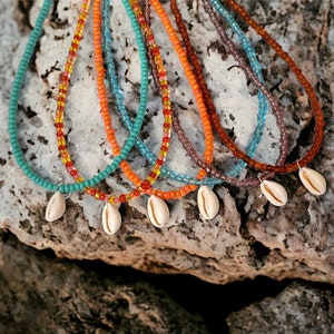 May include: A collection of beaded necklaces in various colors, including teal, orange, and brown. Each necklace features a small, white seashell pendant. The necklaces are displayed on a textured, gray stone surface.